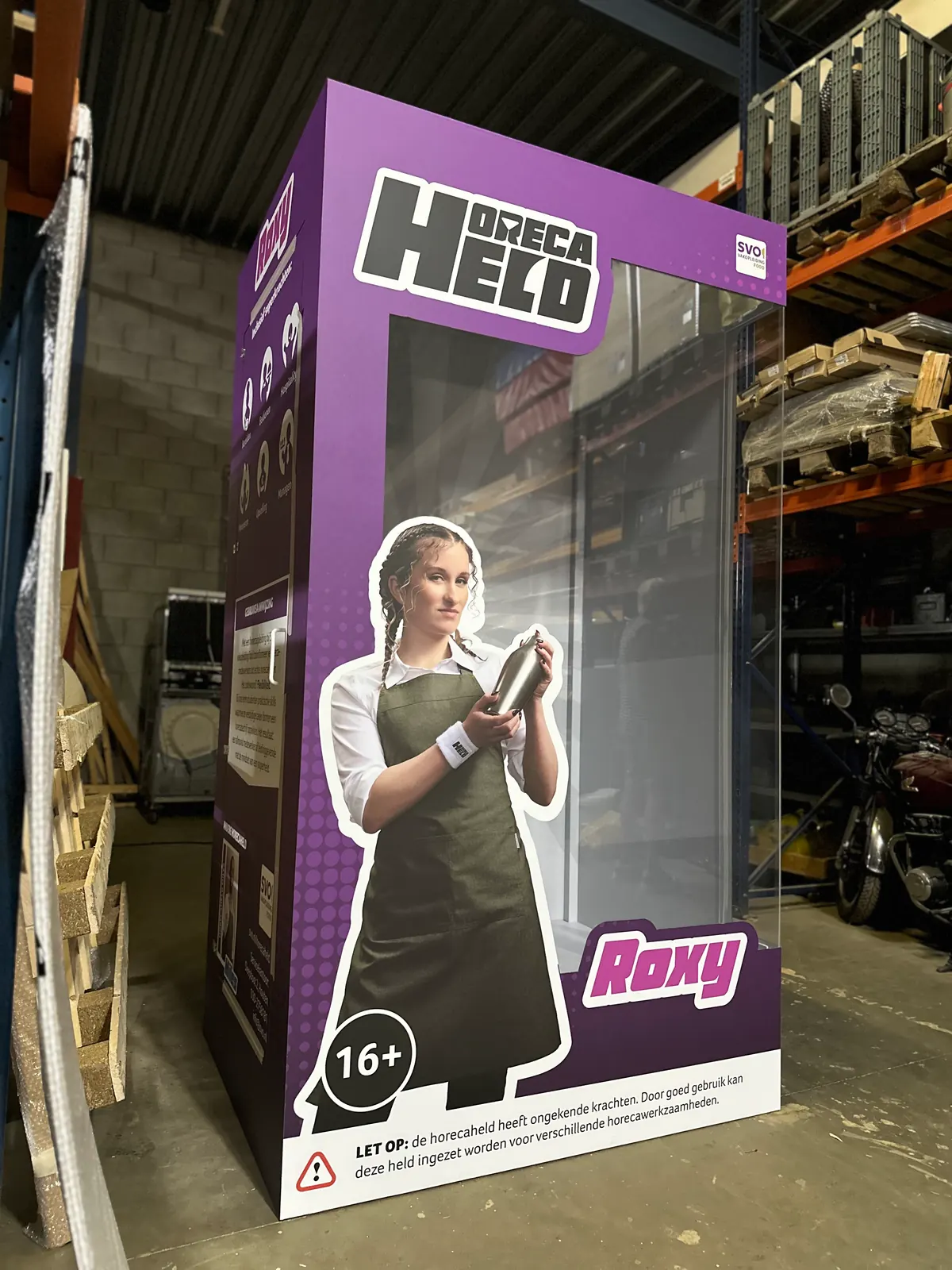 Paarse actieheldendoos op mensformaat met Project Held-opschrift en Roxy-paneel in de werkplaats