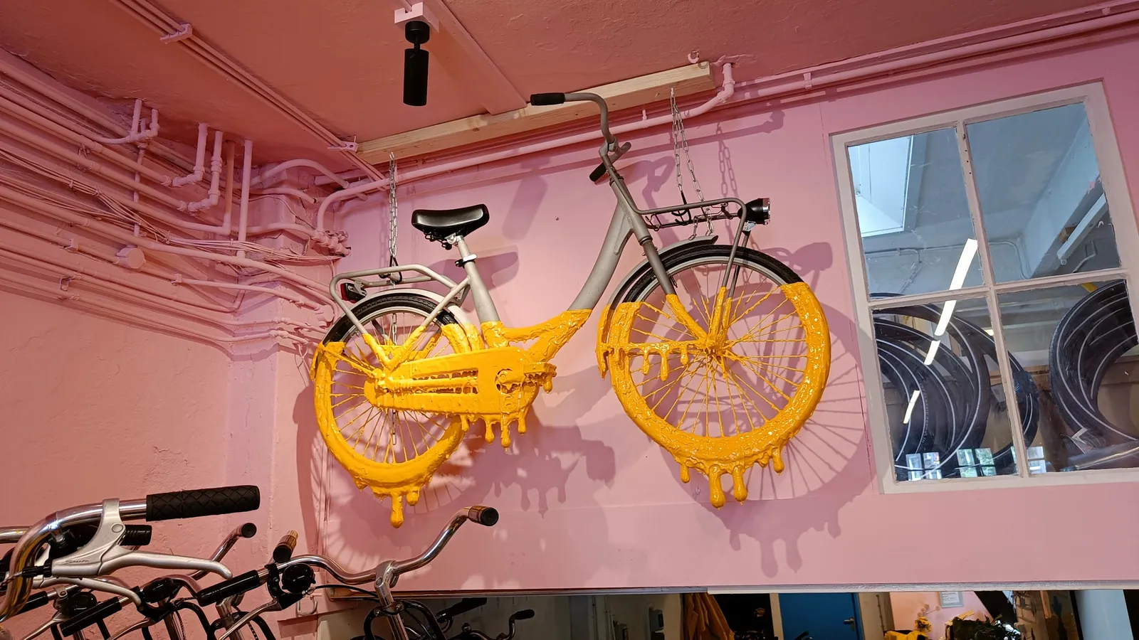 Gele fietssculptuur met druipvormen tegen een roze wand bij Yellow Bike